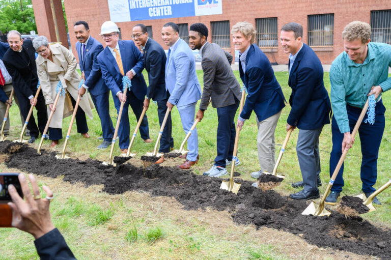 A group of people stand beside each other with shovels in hand as they turn the dirt.